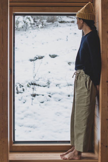 A woman wearing a yellow beanie, blue sweater, and tan pants, all from Ecologyst, stands looking out the window at snow covered ground.