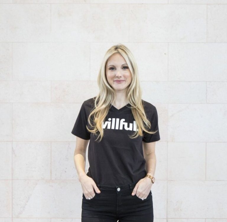 Erin Bury wearing a willful t-shirt and black jeans. She has her hands in her pockets and is standing against a white brick wall.