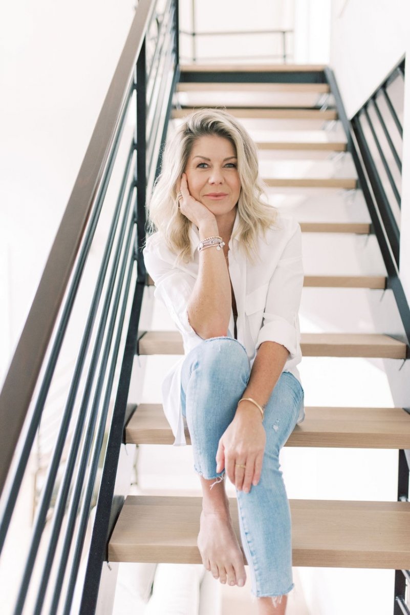 A blonde woman, Janey Reilly, founder of WeeSleep, wearing blue weans and a white shirt sits on wooden stairs. She smiles at the camera.