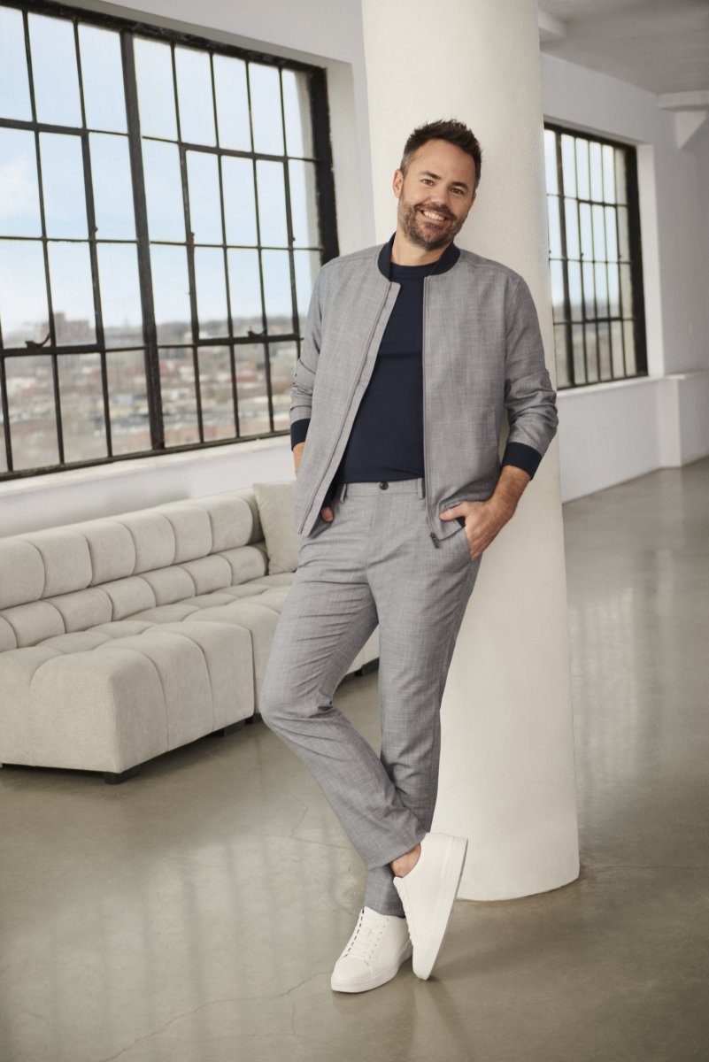 A white man, Lane Merrifield, leans against a wall, wearing a grey bomber jacket and grey pants.