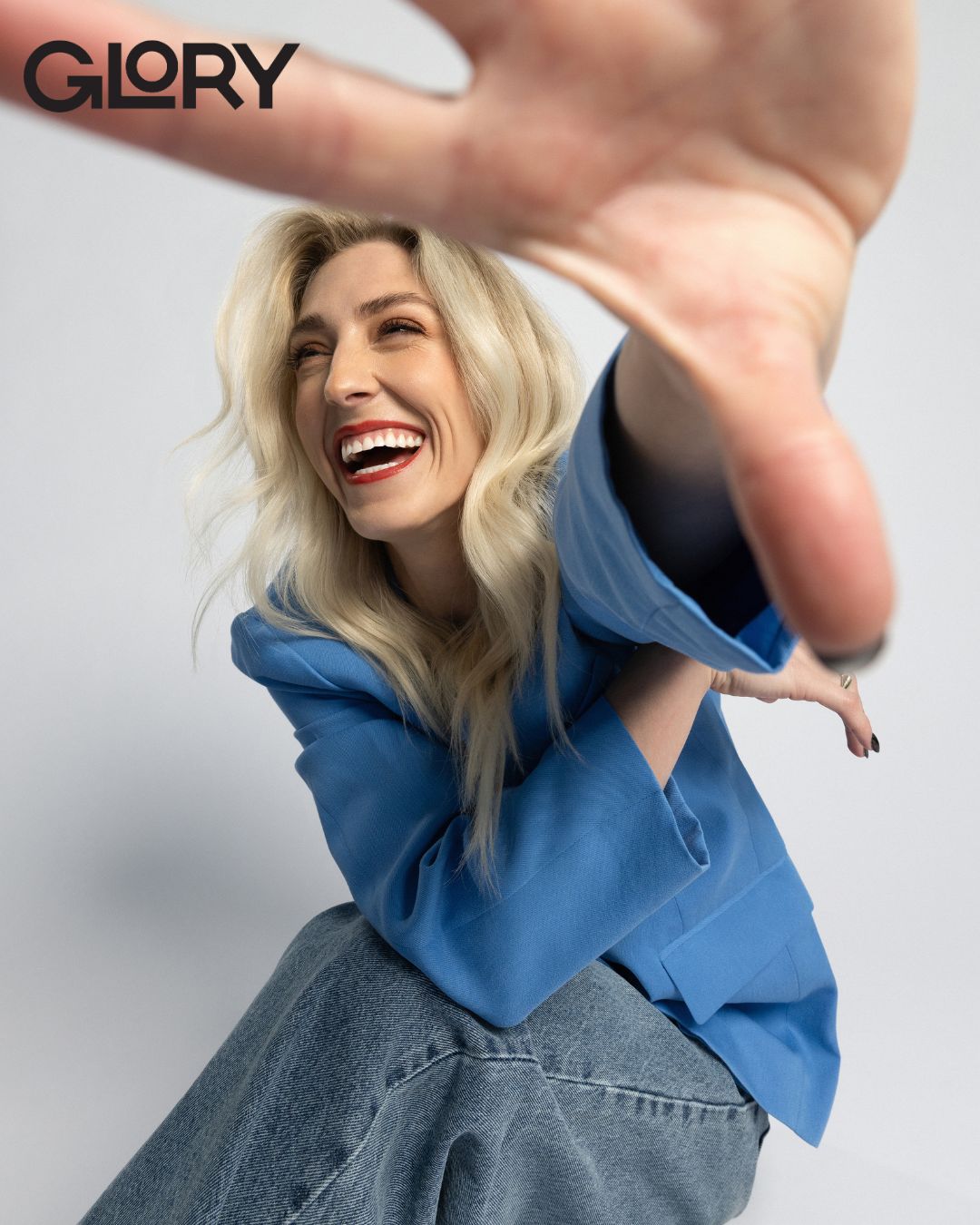 closeup shot of Piper Gilles laughing and a hand reaching out to a camera wearing a blue blazer and jeans.