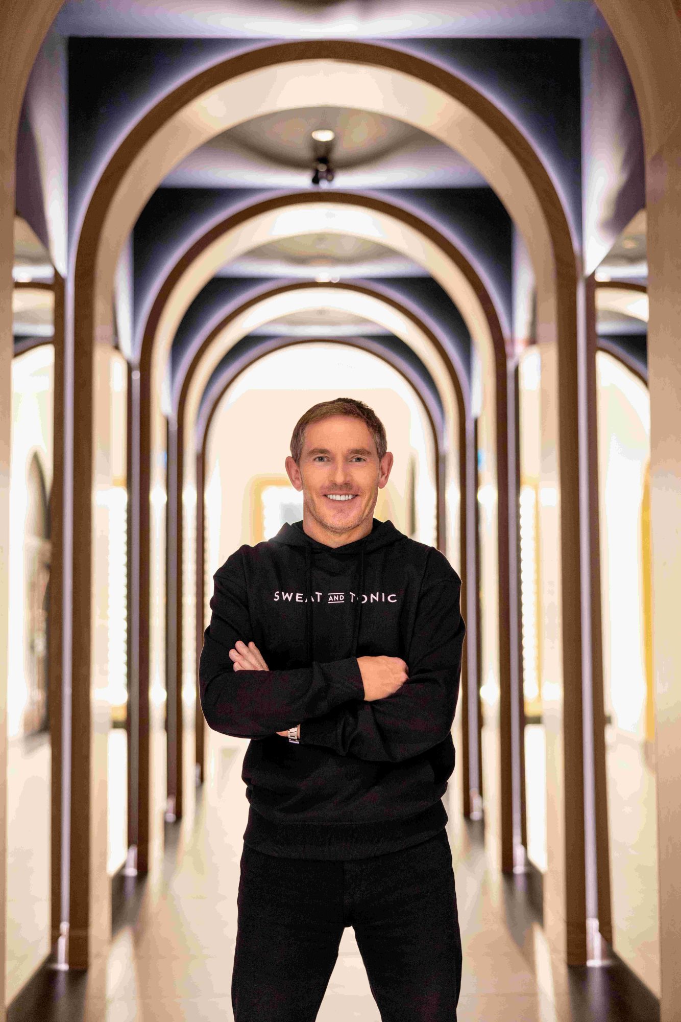 Sweat and Tonic founder David Ingram wearing a branded black hoodie and arms crossed in a hallway.