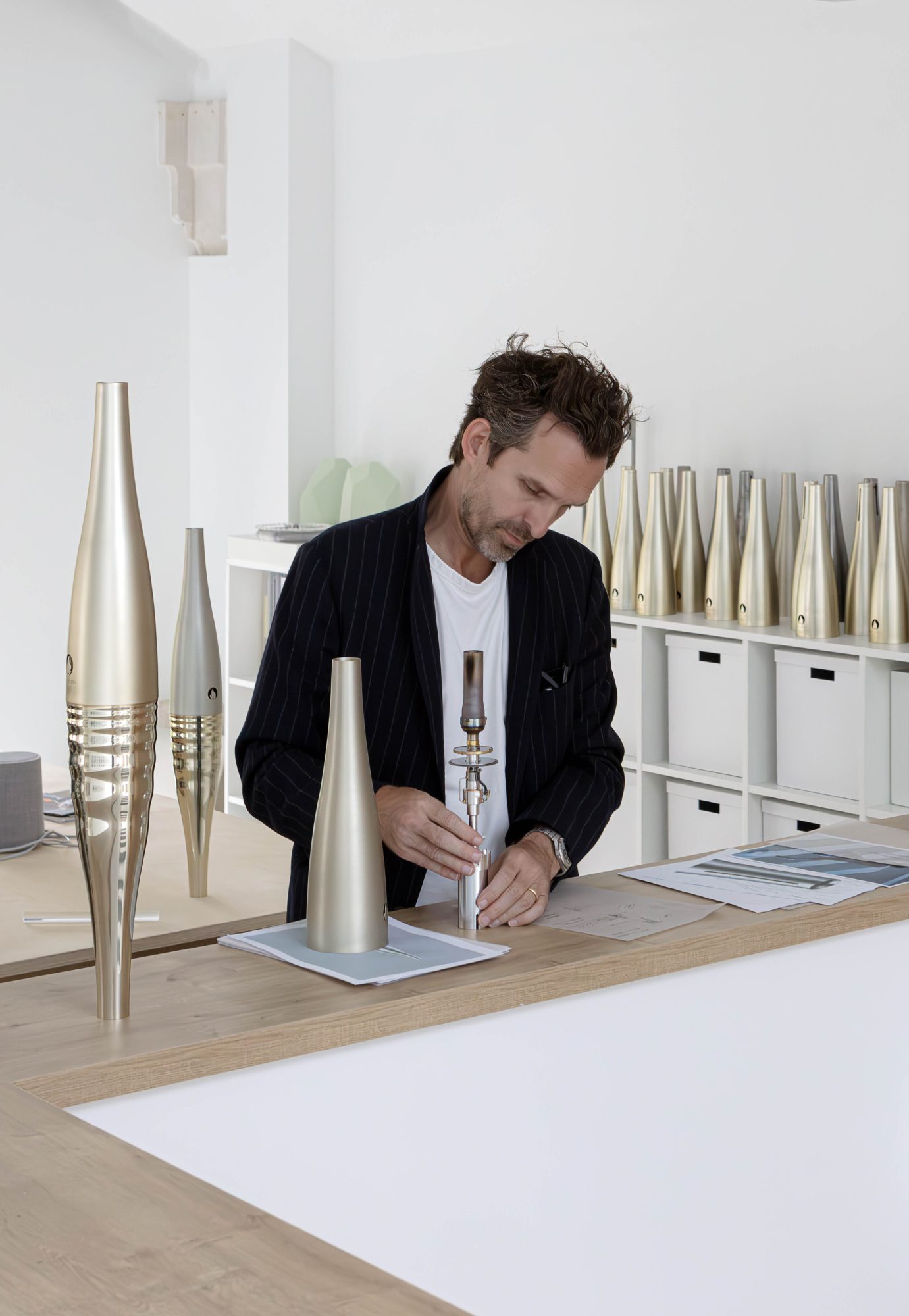 Mathieu Lehanneur in a dark blazer and white t-shirt at a desk with pieces and replicas of the Paris 2024 Olympic Torch