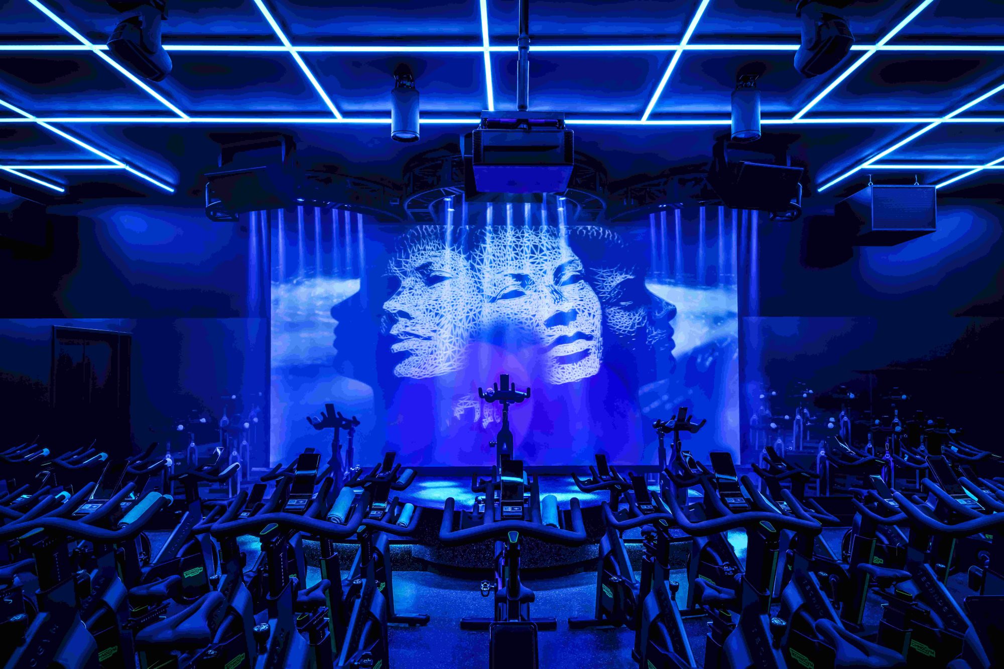 A blue-lit cycling room with the bikes facing a large screen projecting a digital face of a woman.