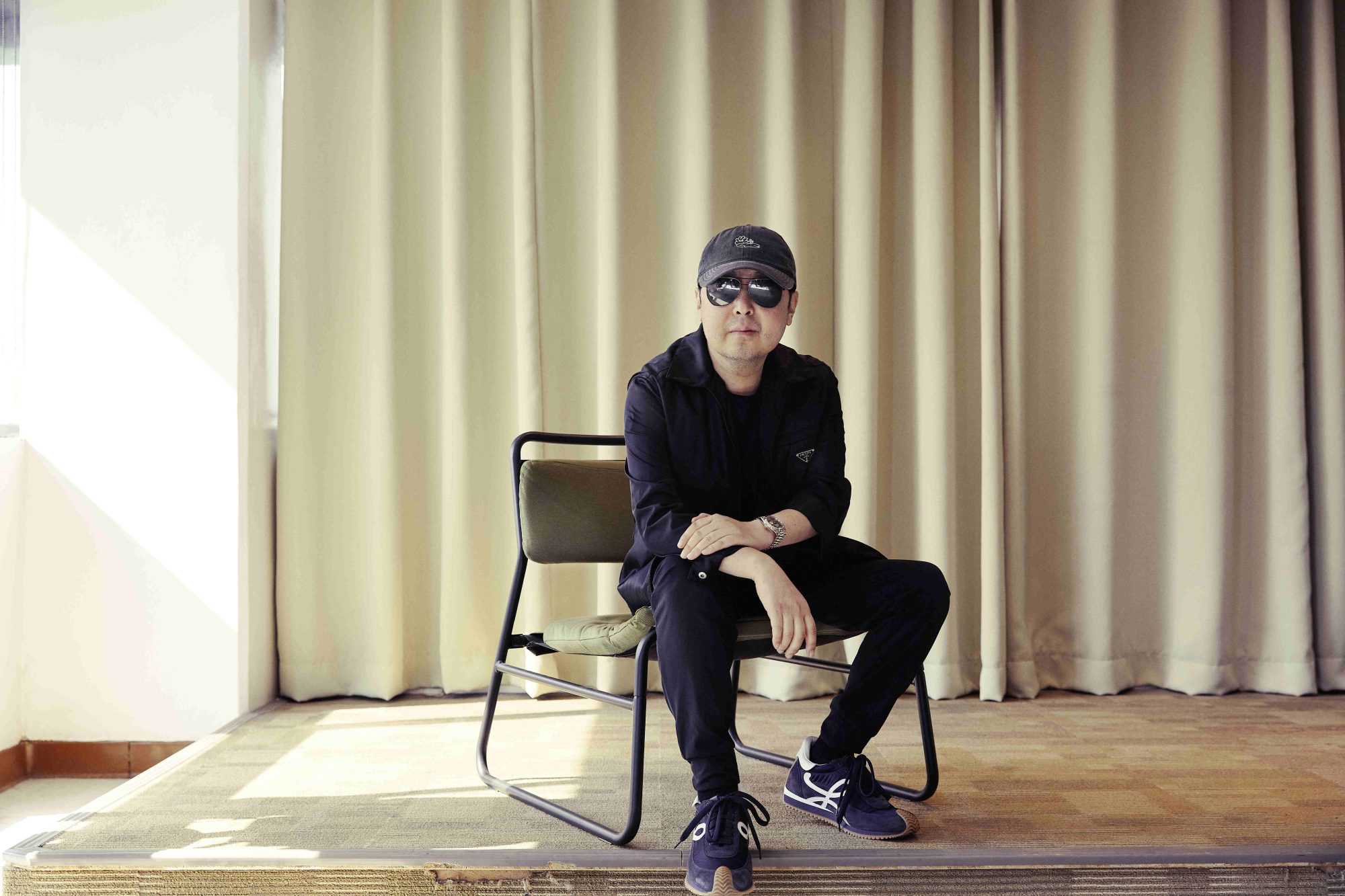 Jia Zhang-Ke wearing all black and seated in a chair against a beige curtain