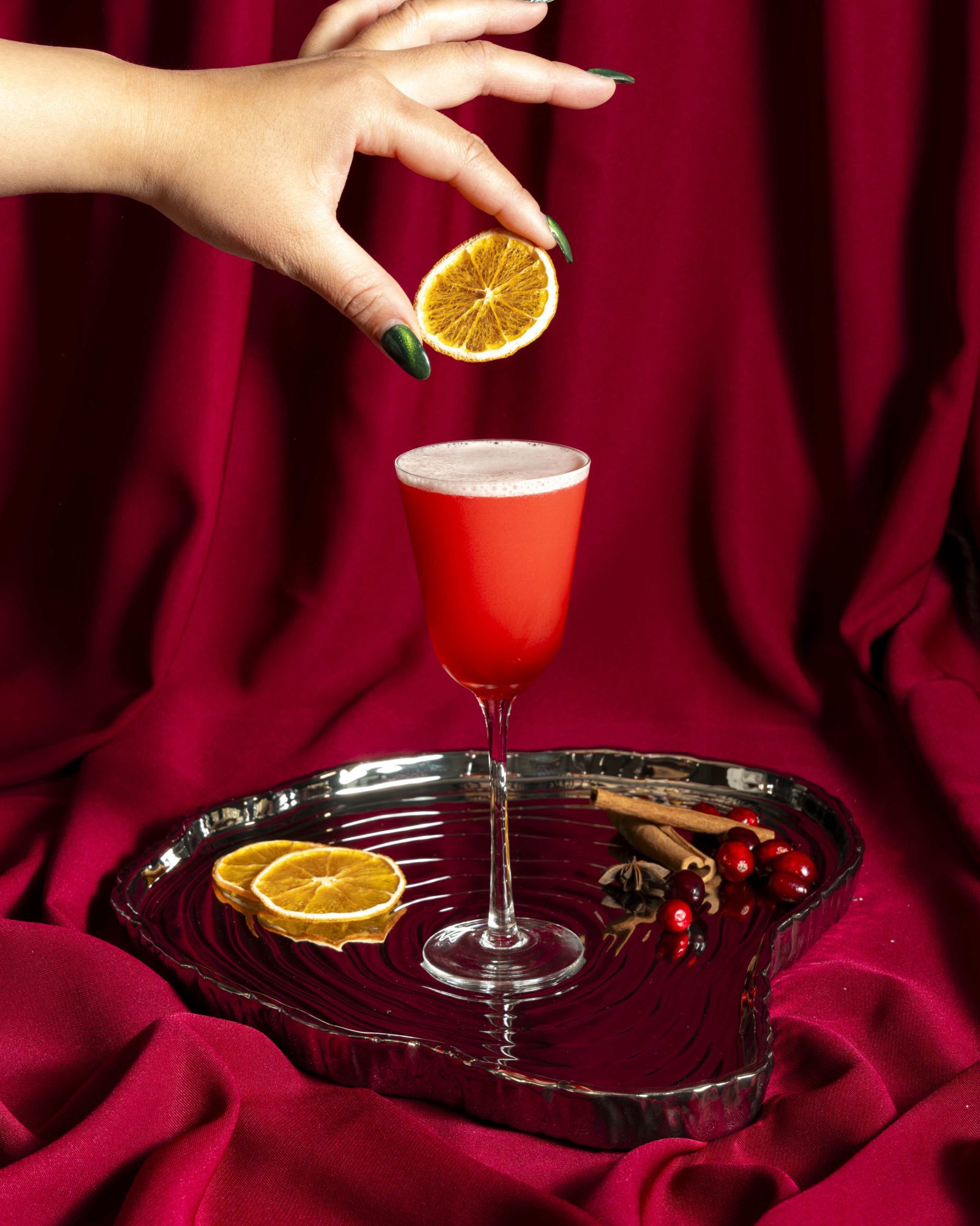 A red cocktail with a hand about to place a dried citrus garnish.