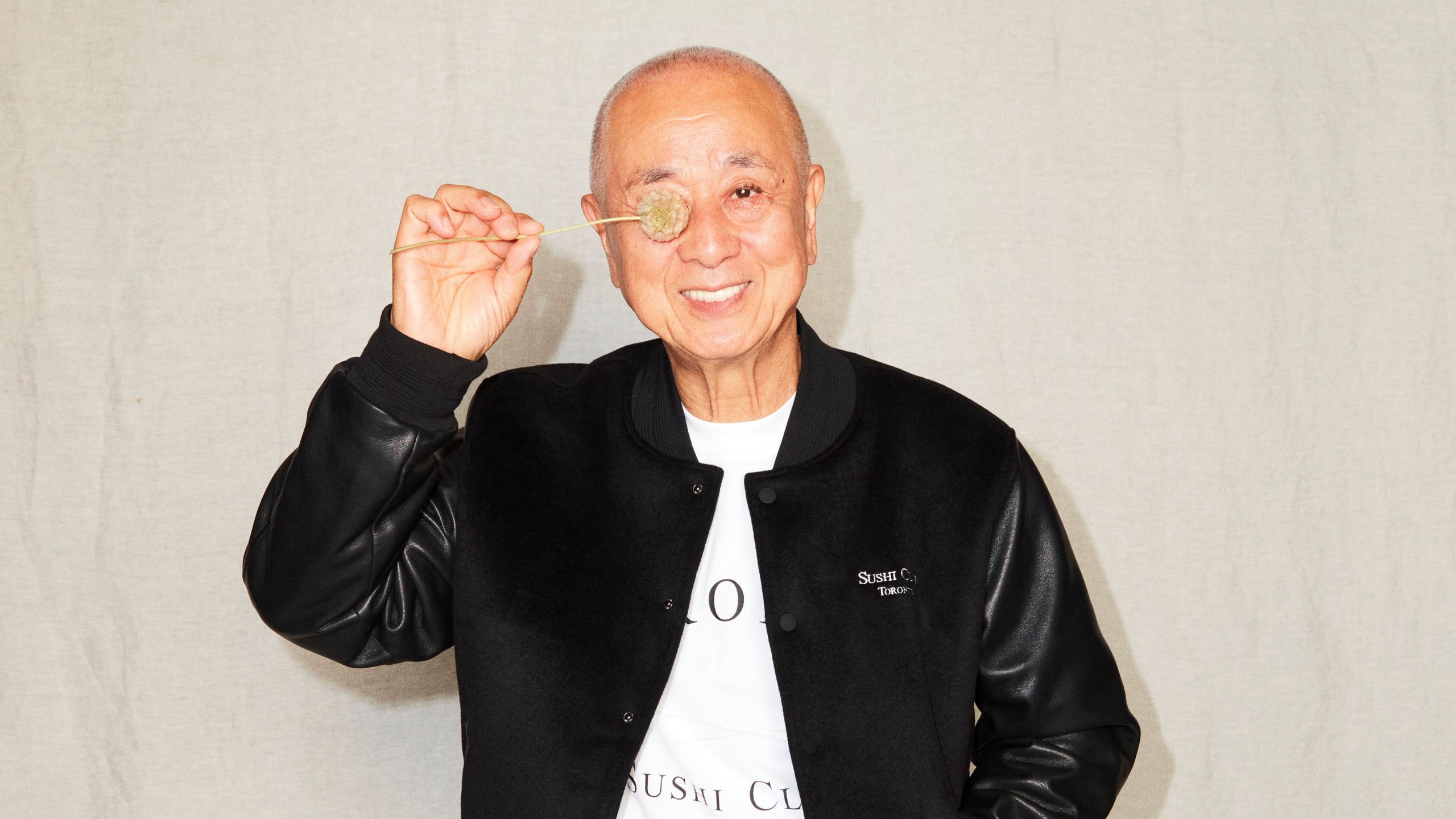 Nobu Matsuhisa holding up a dried flower next to his eye wearing a black jacket.